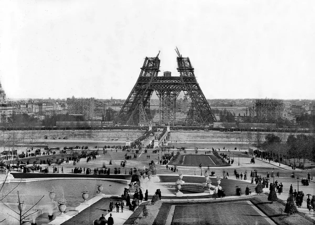 La Tour Eiffel en construction, un aper&ccedil;u de son histoire. Des ouvriers travaillent sur la structure m&eacute;tallique, tandis que des Parisiens se prom&egrave;nent sur le Champ de Mars.
