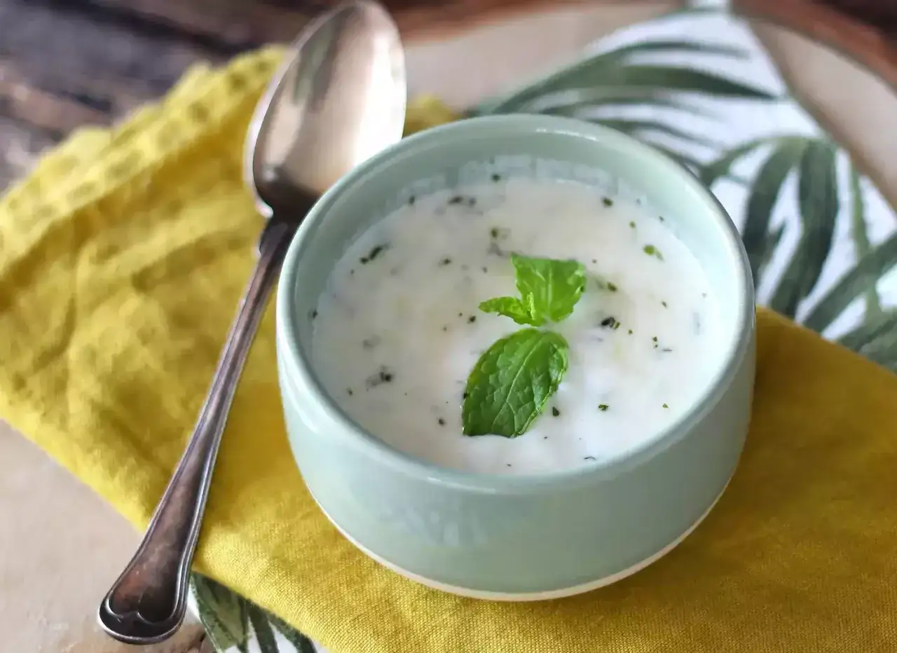 Eine Sch&uuml;ssel cremiger Joghurt-Suppe mit Minze, dane liegt ein L&ouml;ffel auf einem gelben Tuch. Ein k&ouml;stlicher Fisch mit Joghurt-Dressing wartet.