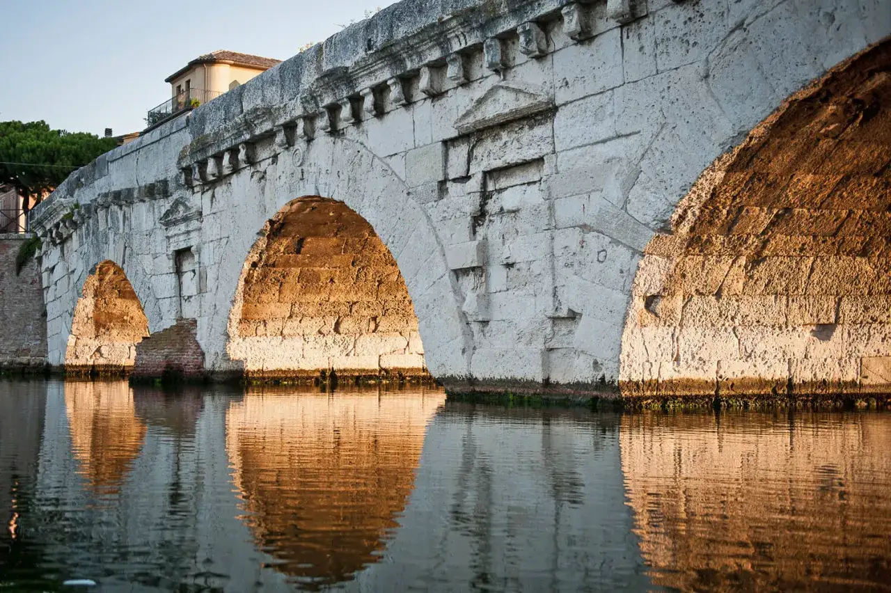 Il Ponte di Tiberio a Rimini, con i suoi archi in pietra bianca illuminati dal sole dorato, si specchia sull'acqua calma.