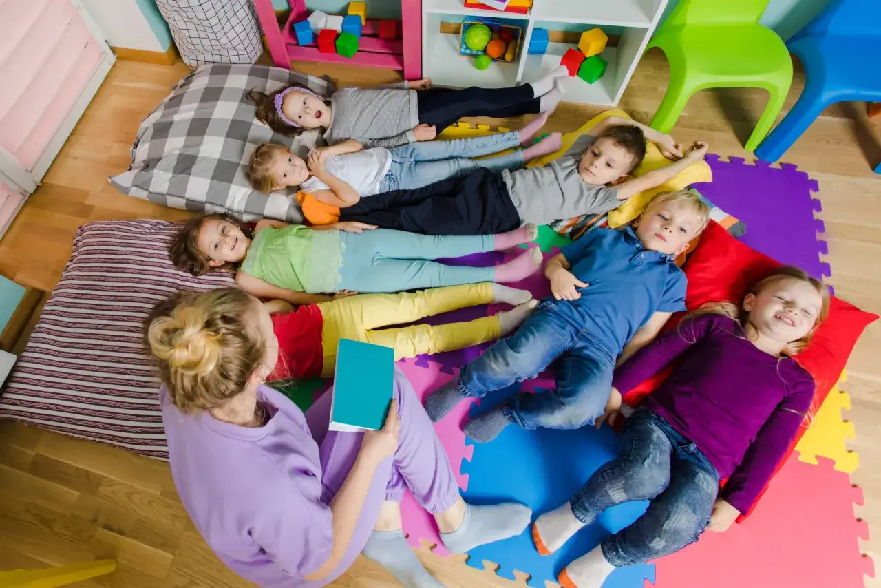 Kinder liegen auf bunten Matten, bereit für eine Geschichte. Die Lehrerin liest vor, wann kein Mittagsschlaf mehr ist.
