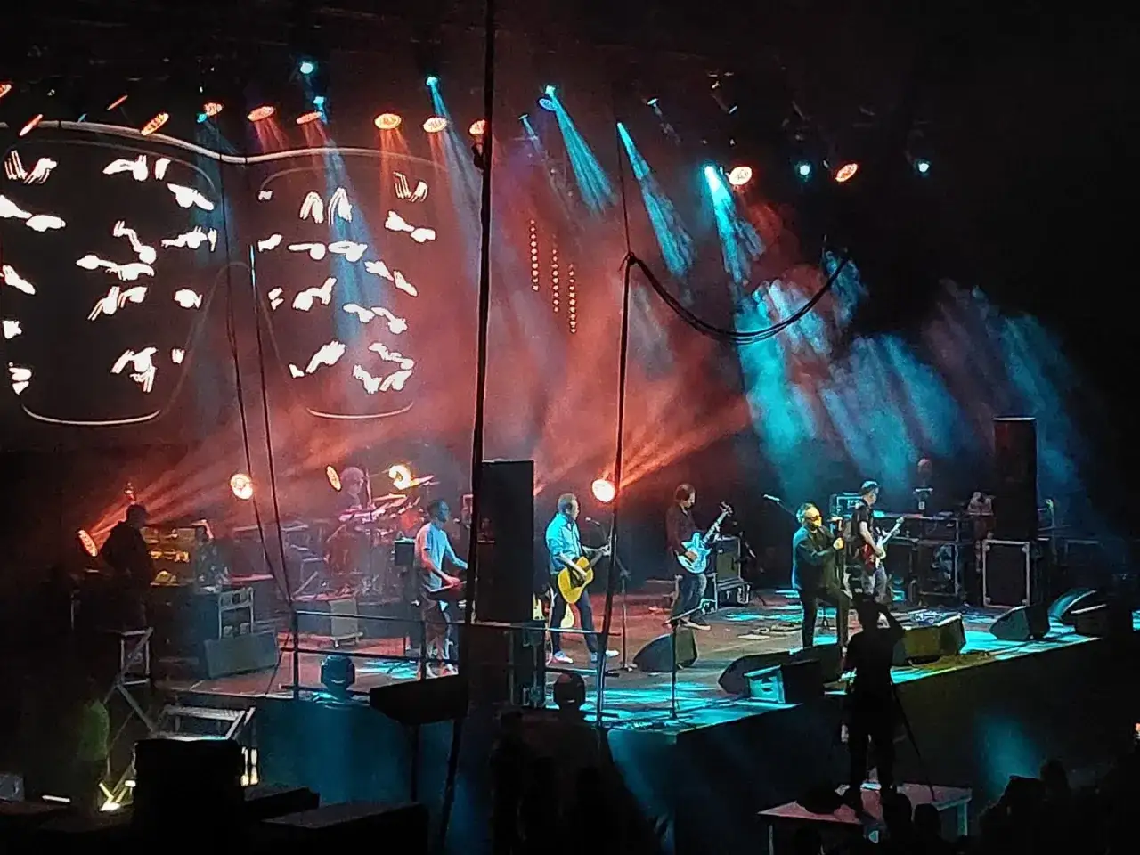 Zespół T.Love na scenie, w otoczeniu dymu i kolorowych świateł. Muzycy grają na gitarach, perkusji, tworząc energetyczny koncert.