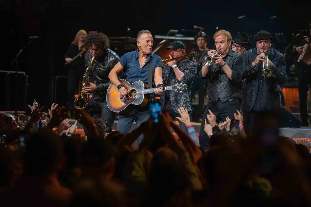 Bruce Springsteen z gitarą i zespołem na scenie podczas jednego z jego niezapomnianych koncertów. Fani w tłumie wznoszą ręce.
