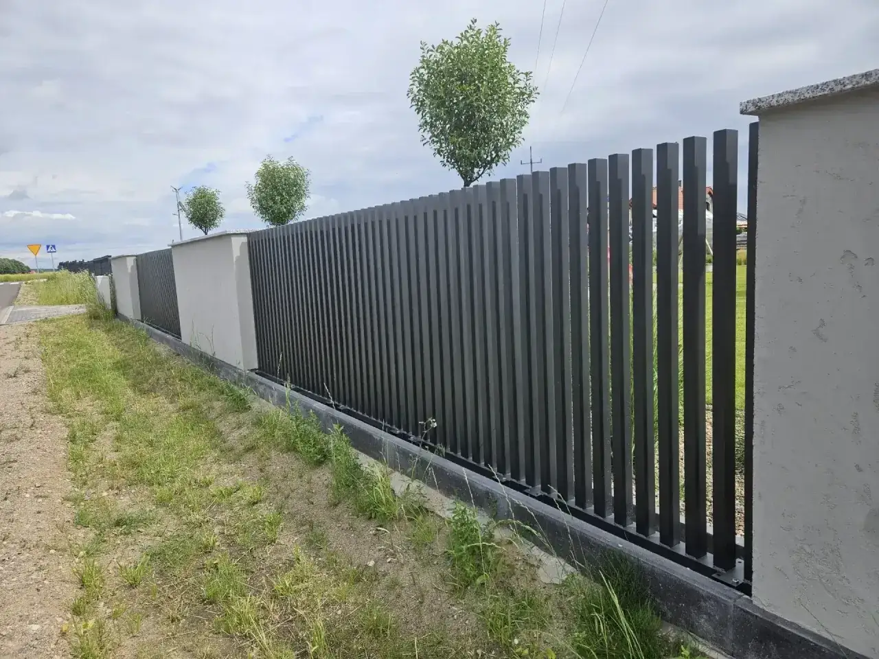 Nowoczesne ogrodzenie na skarpie, z pionowych, ciemnych paneli i betonowych słupk&oacute;w, otacza zielony teren.