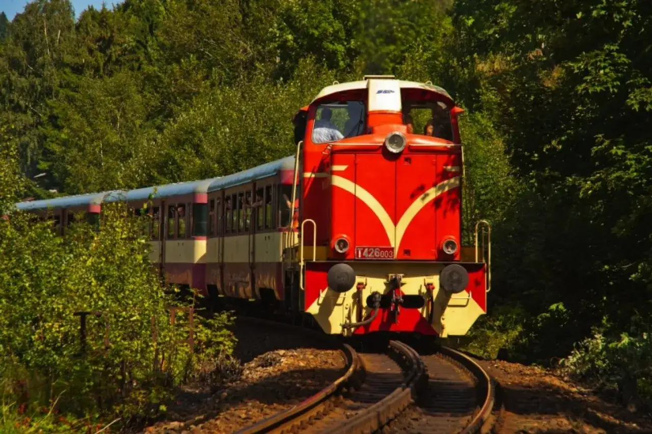 Czerwony pociąg z żółtymi pasami jedzie przez zielony las. To malownicza trasa pociąg Szklarska Poręba Harrachov.