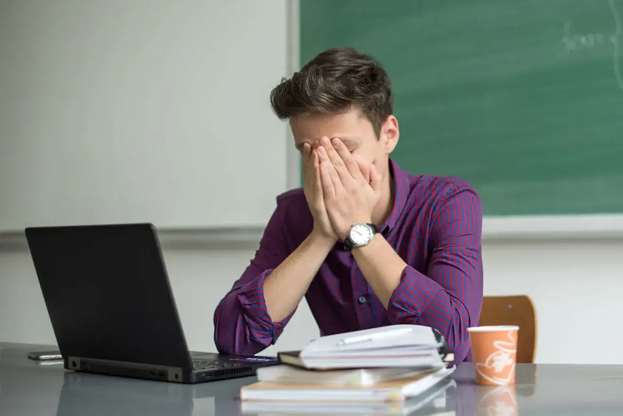 A young man with his face in his hands, contemplating how to recover from a mistake at work. A laptop and stack of books are on his desk.