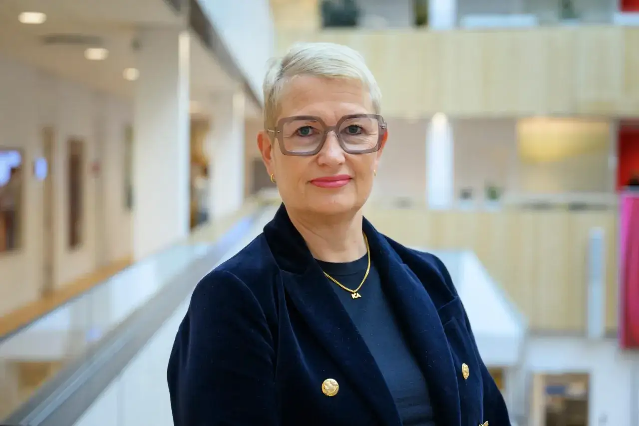 En kvinna med glasögon och kort blont hår, klädd i en mörkblå kavaj, bär ett halsband med bokstäverna