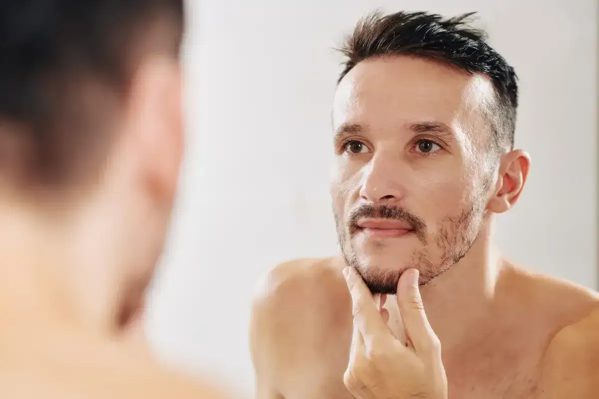 Mężczyzna w lustrze zastanawia się, jak pobudzić zarost na twarzy. Delikatnie dotyka swojej brody, szukając inspiracji.