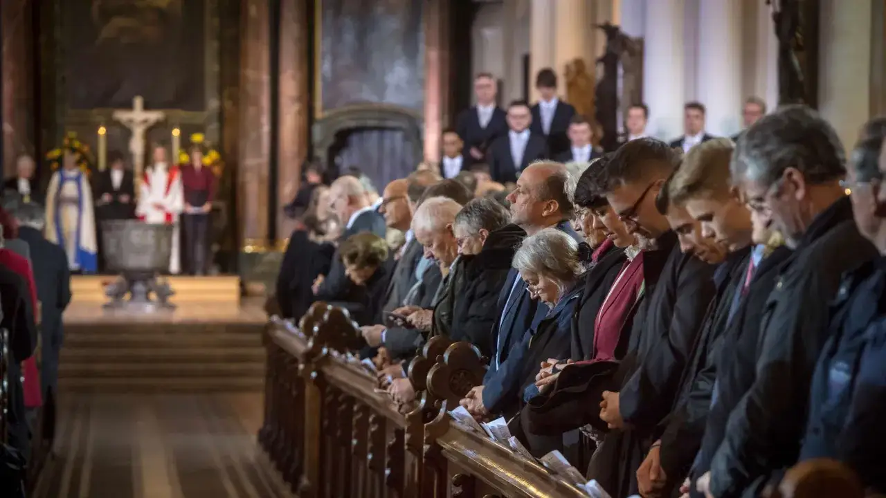 Menschen in einer Kirche, die gemeinsam das Glaubensbekenntnis sprechen. Im Vordergrund sitzen Gl&auml;ubige auf Kirchenb&auml;nken, im Hintergrund ist der Altarbereich zu sehen.