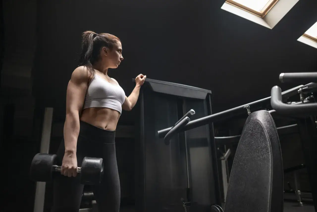 En kvinna med en rippad kropp h&aring;ller en hantel i ett m&ouml;rkt gym.