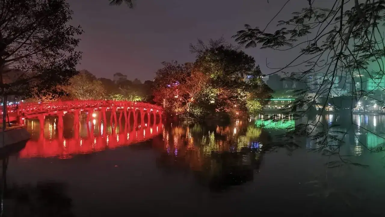 Czerwony most w Hanoi, Wietnam, odbija się w wodzie nocą. Drzewa oświetlone kolorowymi światłami.