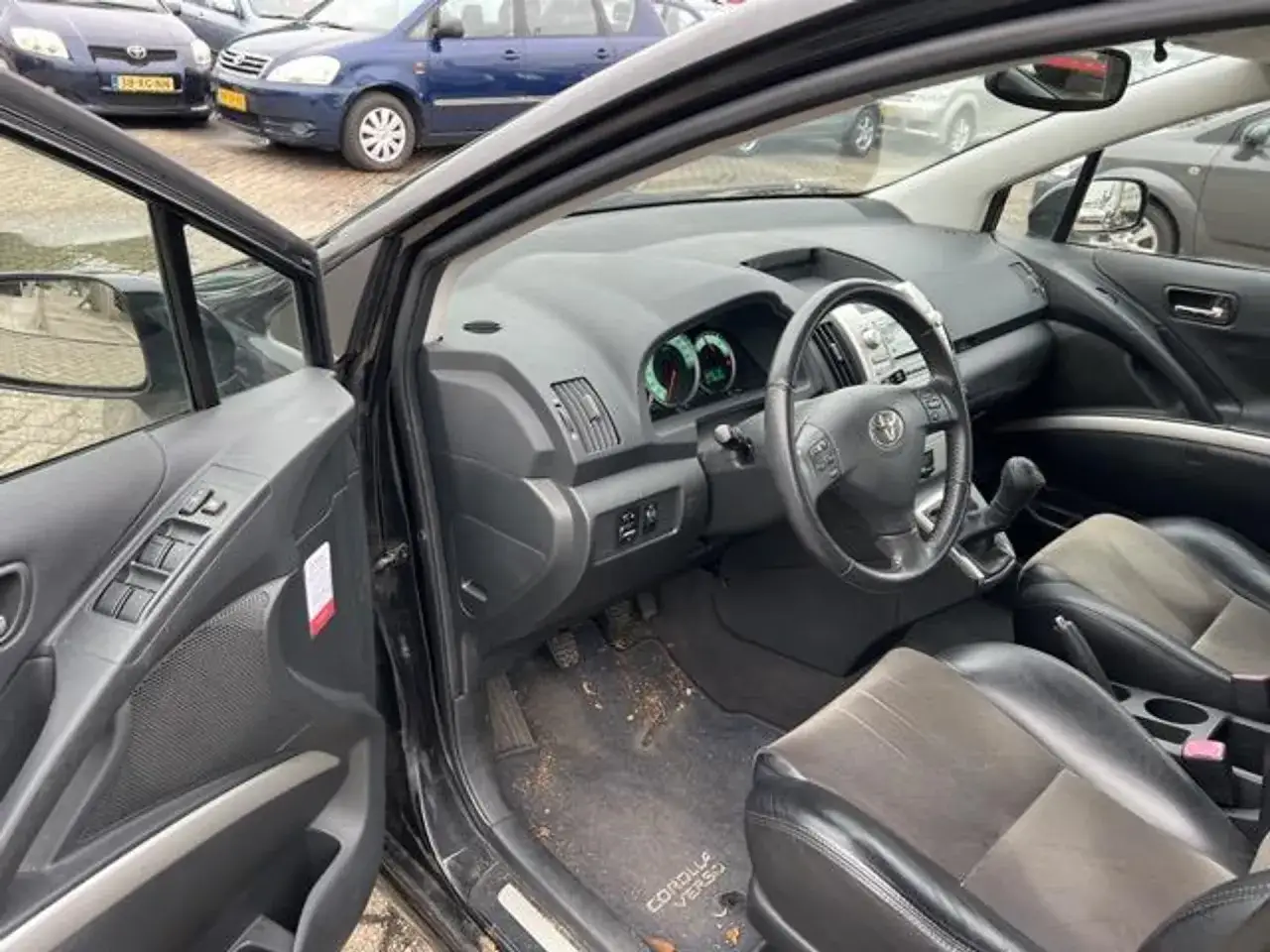 Wnętrze Toyoty Corolla Verso z widoczną kierownicą, deską rozdzielczą i siedzeniami. Na dywaniku widoczny napis 
