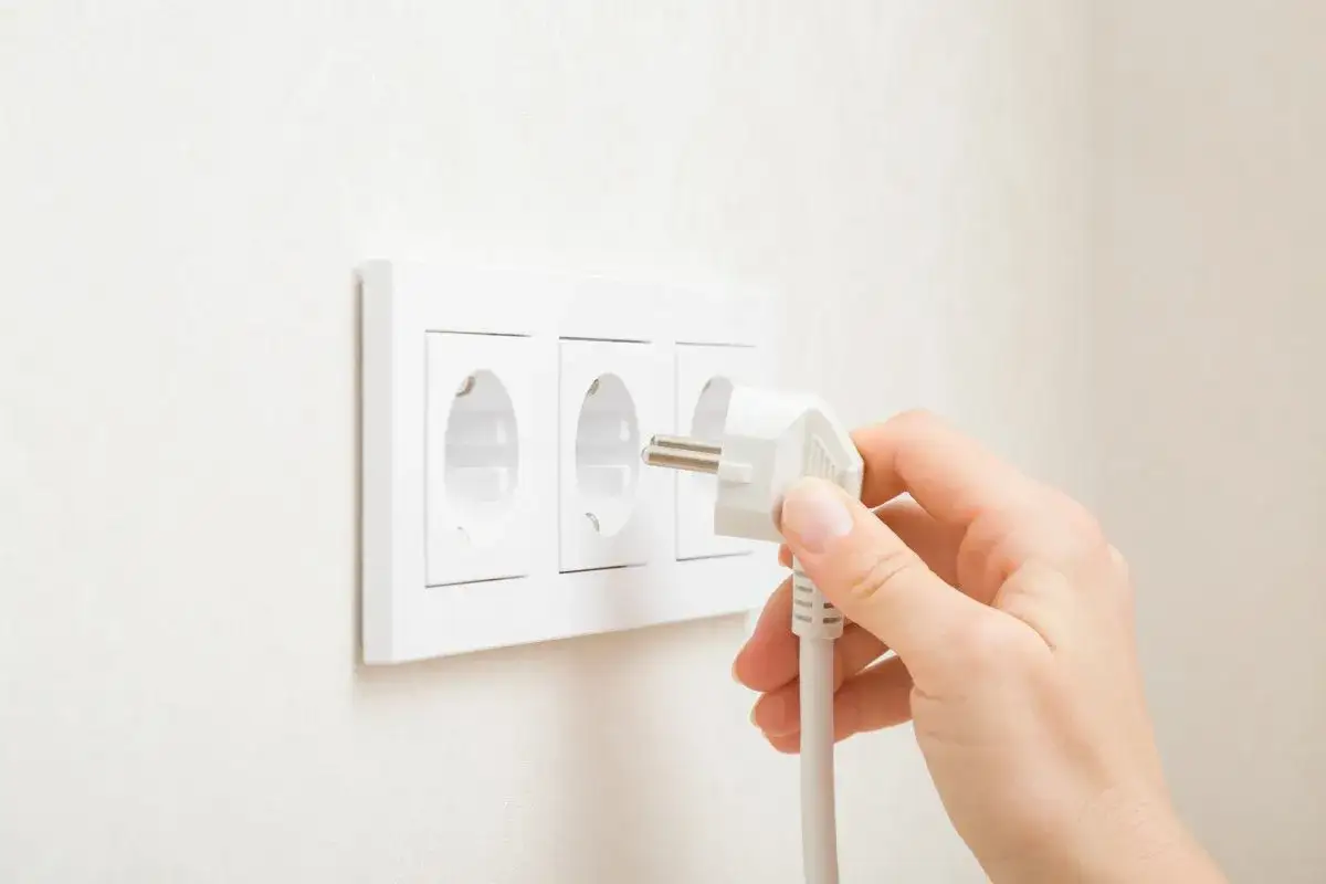 Main d'une personne insérant une prise dans une prise électrique murale triple. La prise est blanche, avec des broches métalliques visibles. La prise murale est également blanche et fixée sur un mur texturé. La scène évoque la connexion pour la puissan...
