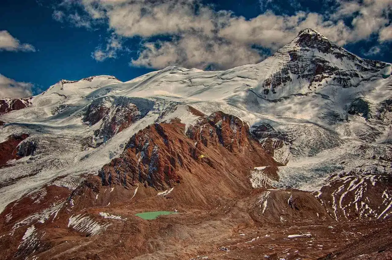 Majestatyczny Aconcagua szczyt w Andach, pokryty śniegiem i lodem, z czerwonymi skałami i turkusowym jeziorkiem u podn&oacute;ża.
