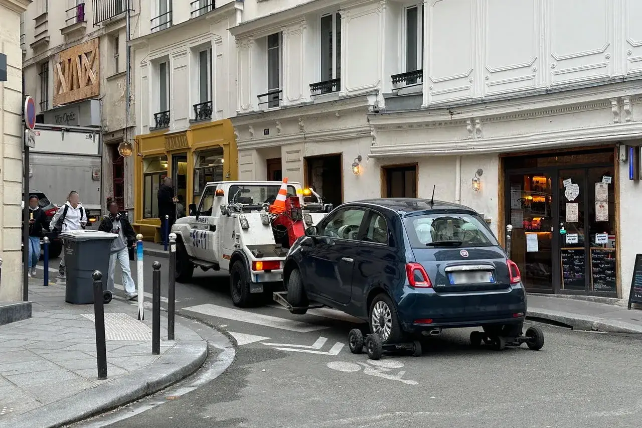 Une dépanneuse enlève une petite voiture bleue. Si vous vous demandez comment savoir si ma voiture est à la fourrière, regardez si elle a disparu de sa place de stationnement.