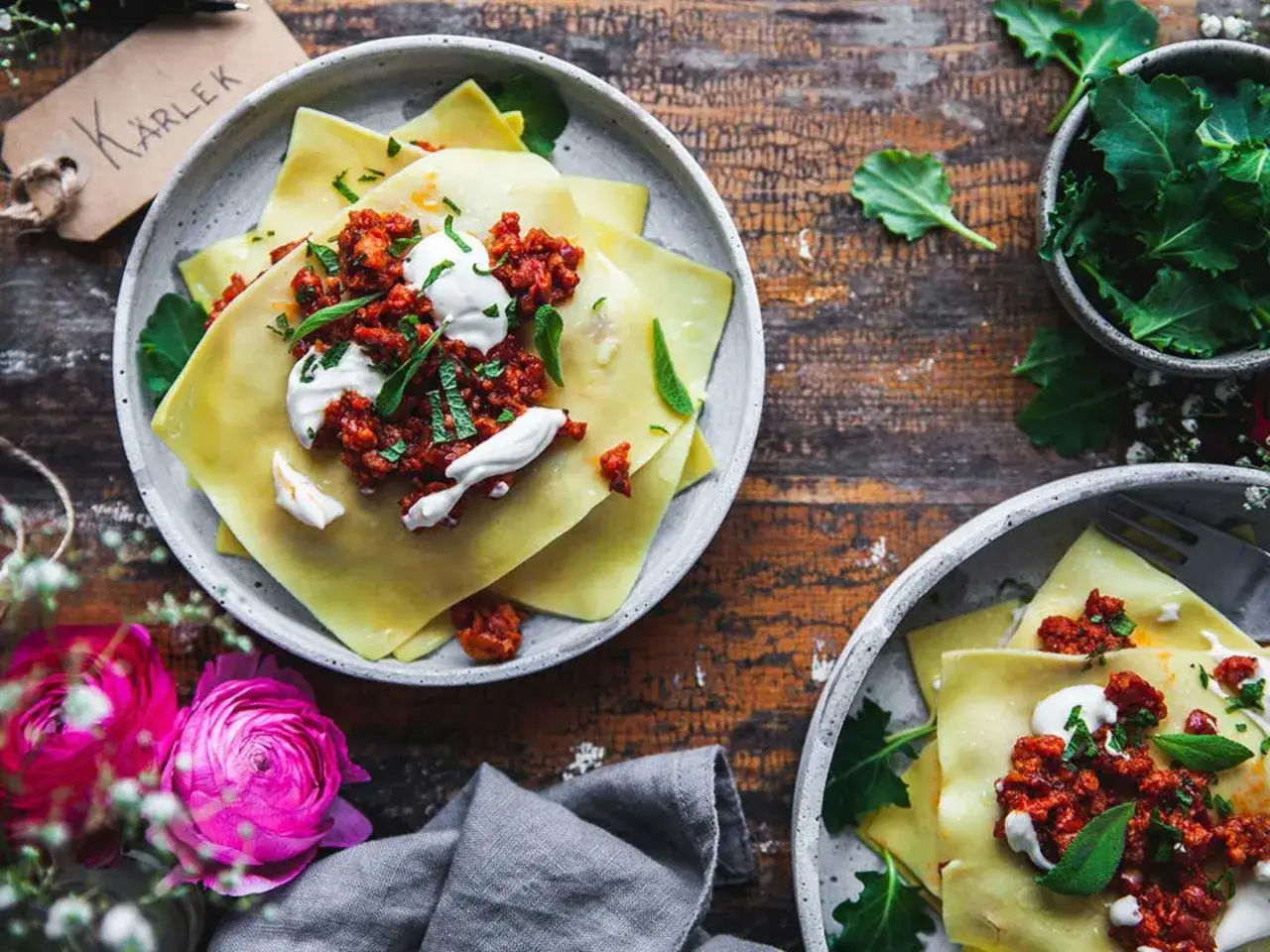 Två tallrikar med lasagneplattor toppade med köttfärssås och gräddfil. En skål med gröna blad och en tagg med texten