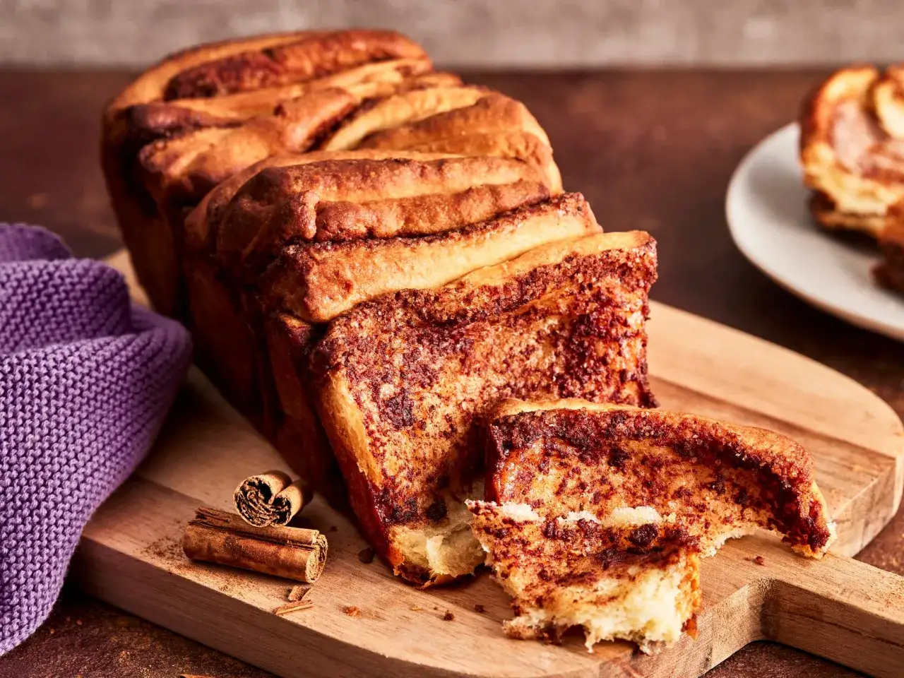 Ein goldbraunes, duftendes Monkey Bread mit Zimt und Zucker, auf einem Holzbrett, daneben Zimtstangen und eine lila Strickdecke.