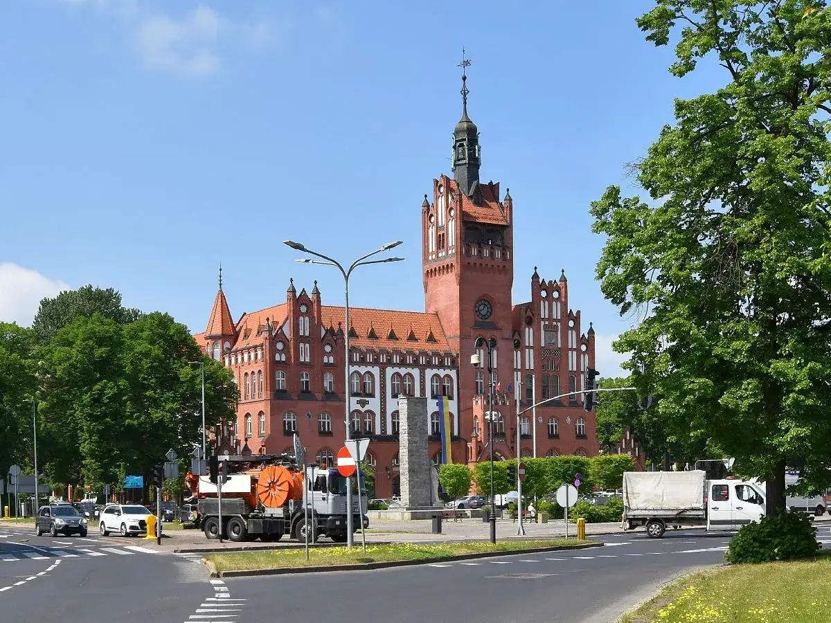Majestatyczny ratusz Słupsk z czerwoną cegłą i wieżą, otoczony zielenią. Na pierwszym planie ruch uliczny.
