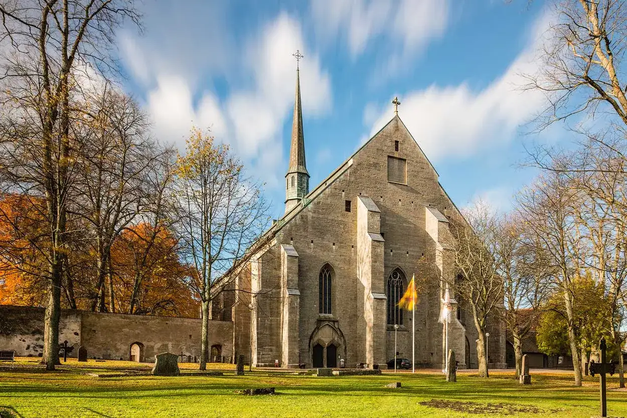 Den heliga Birgittas klosterkyrka i Vadstena, en imponerande stenbyggnad med ett h&ouml;gt torn, omgiven av h&ouml;stf&auml;rgad natur och en kyrkog&aring;rd.