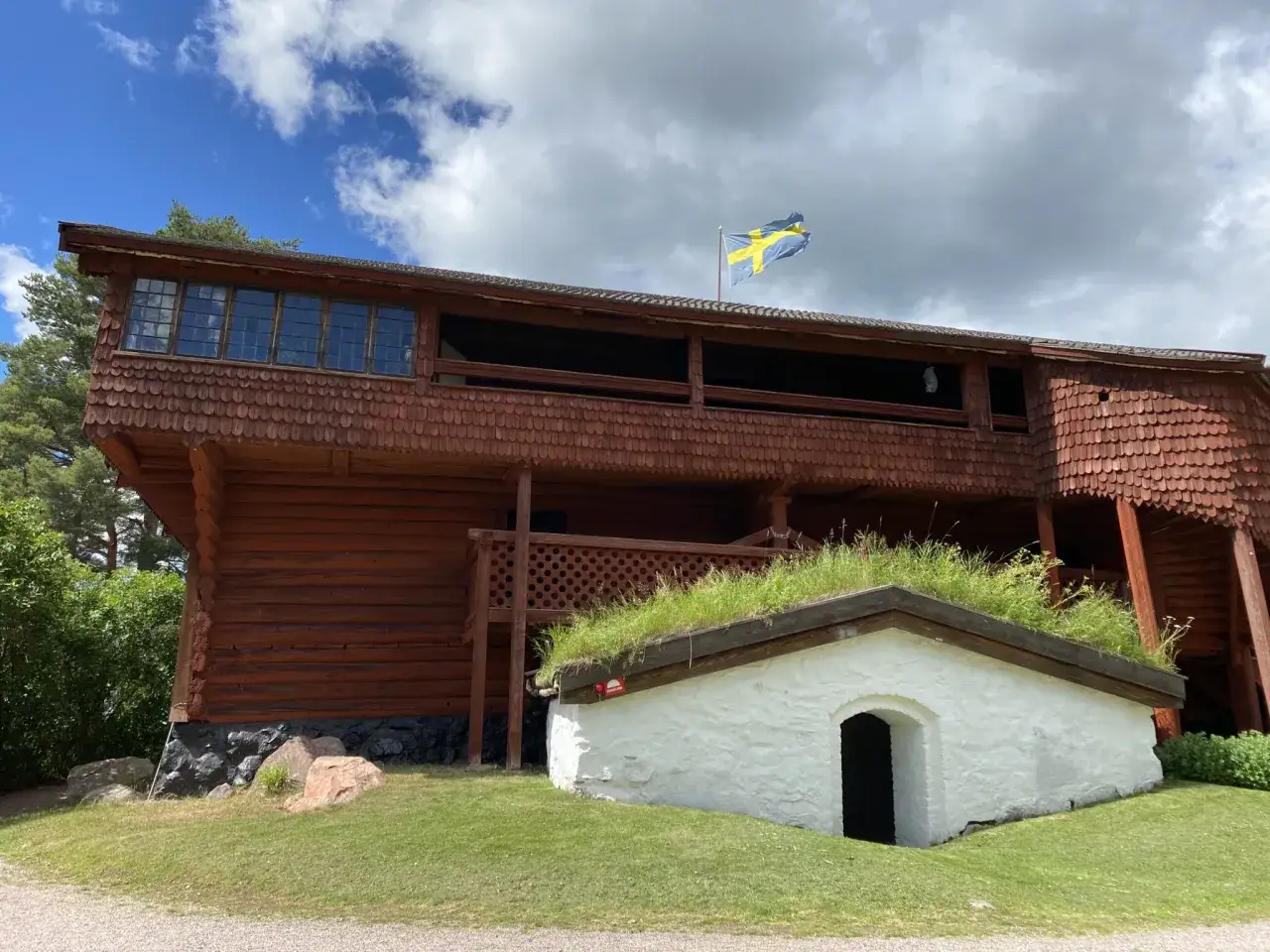 En tr&auml;byggnad med svensk flagga, som fr&aring;n en vasatiden film. Ett litet vitt hus med gr&auml;stak syns framf&ouml;r.