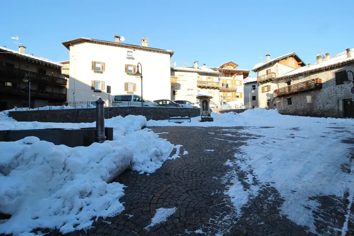 Piazza innevata a Miola di Pinè, con fontana e case tradizionali.
