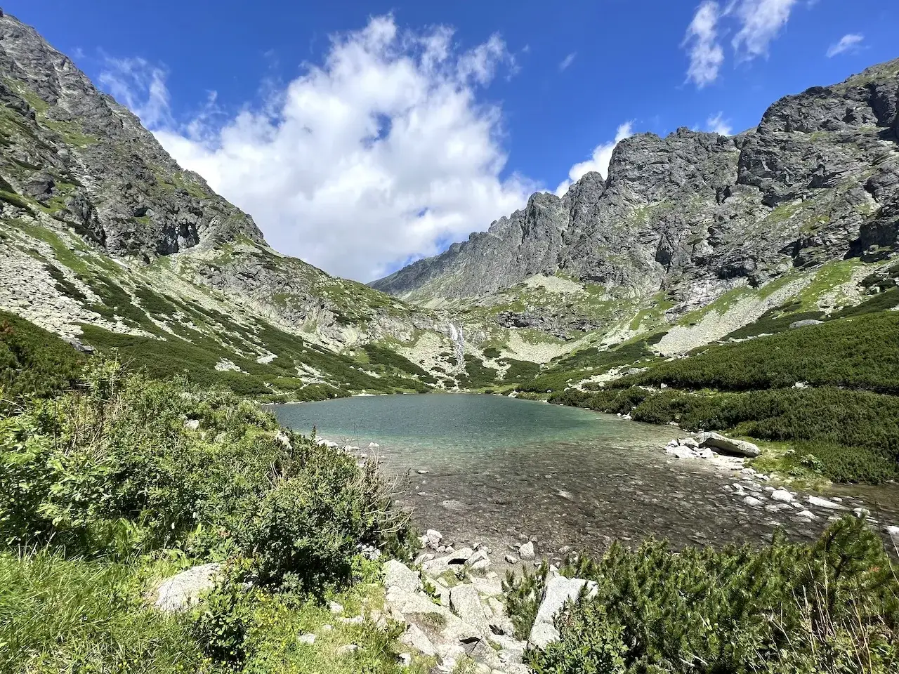 Klares Bergseeufer in den Hohen Tatra, umgeben von steilen, gr&uuml;nen H&auml;ngen und felsigen Gipfeln unter blauem Himmel mit Wolken.