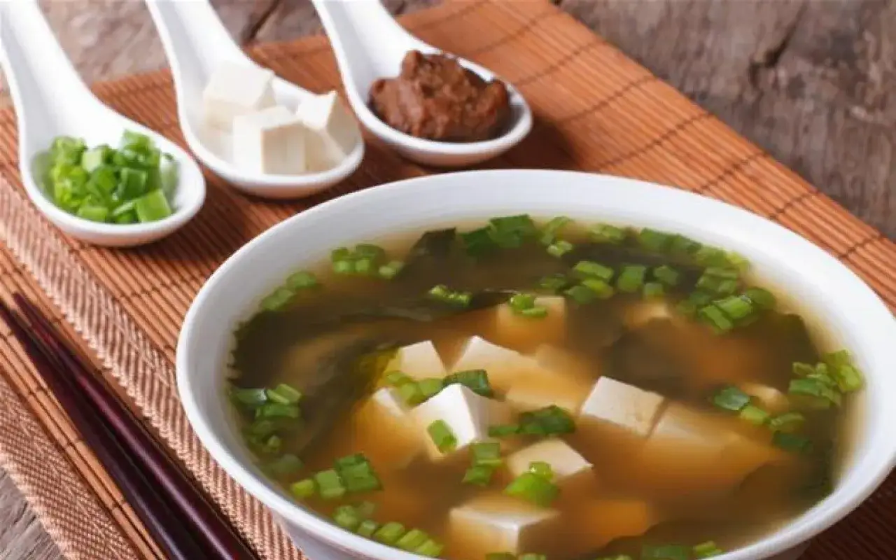Un bol de soupe miso avec des cubes de tofu, des algues et des oignons verts cisel&eacute;s. Trois cuill&egrave;res pr&eacute;sentent des ingr&eacute;dients : oignons verts, tofu et p&acirc;te miso.