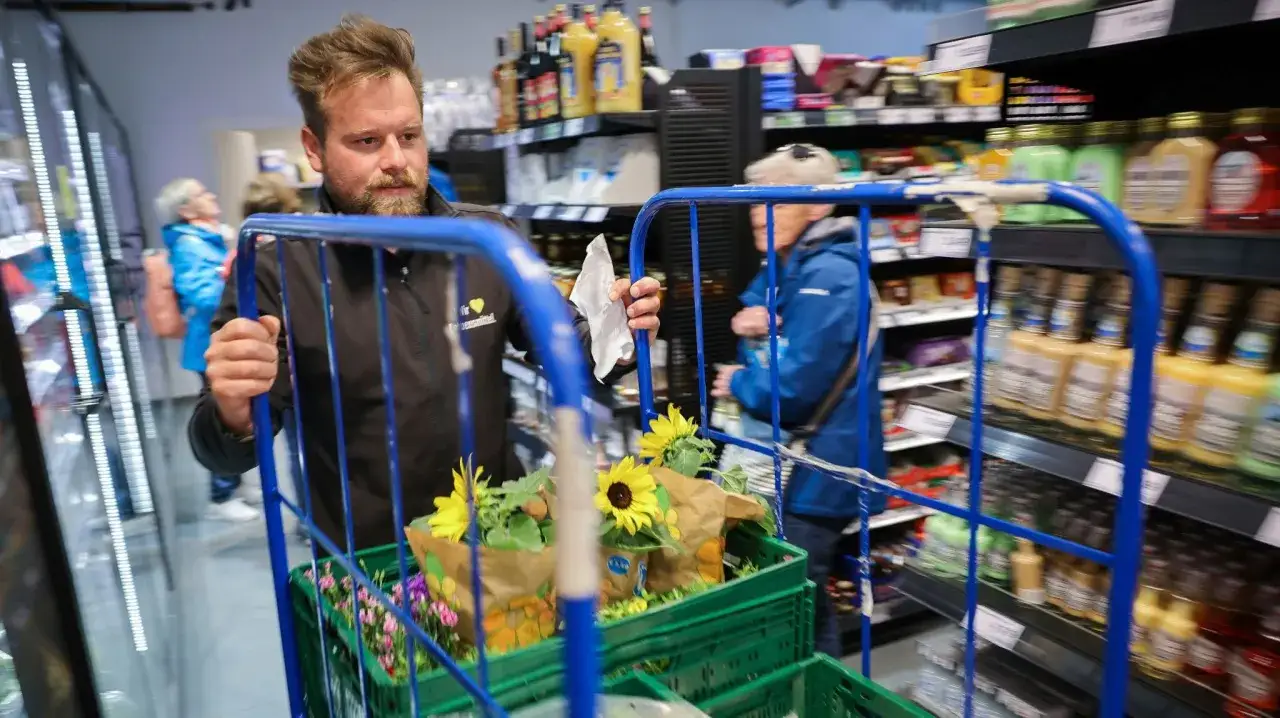 Mitarbeiter schiebt Einkaufswagen mit Blumen und Pflanzen. Hinter ihm Kunden im Edeka. Streik Edeka heute?