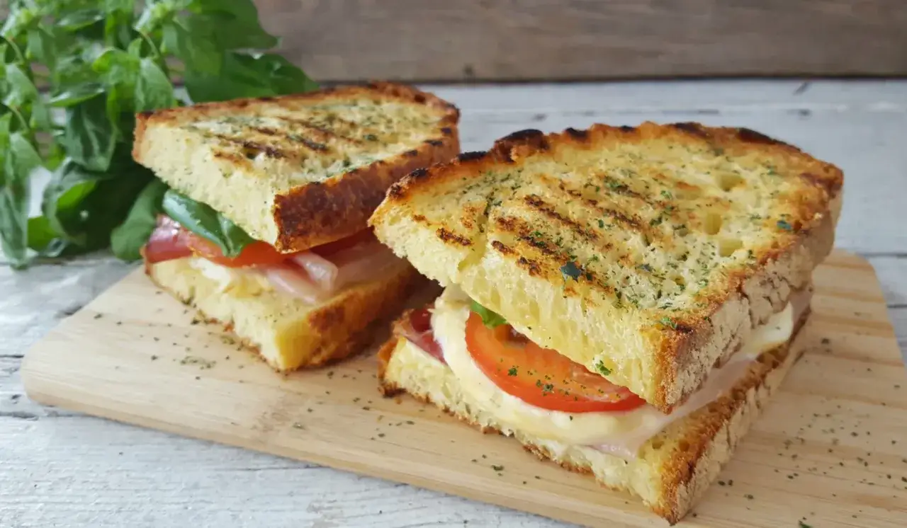 Due panini alla zingara, con pane grigliato, pomodoro, mozzarella e prosciutto, su un tagliere di legno.