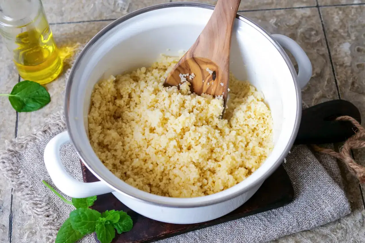 Ein Holzl&ouml;ffel lockert den frisch zubereiteten Couscous in einem Topf. Daneben stehen &Ouml;l und Minze.