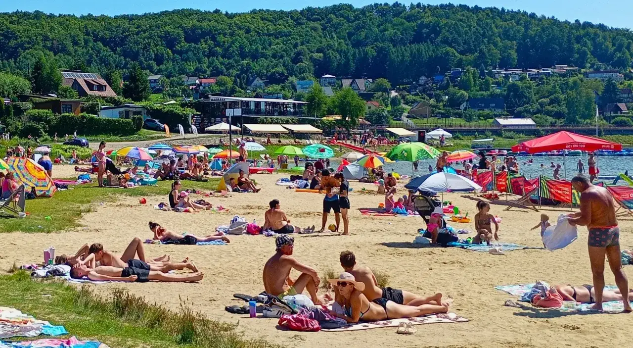 Tłoczna **jezioro rożnowskie plaża** w słoneczny dzień. Ludzie opalają się, kąpią i odpoczywają pod kolorowymi parasolami.