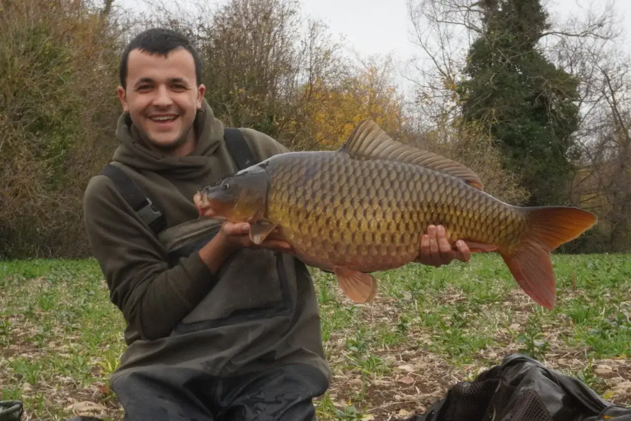 Un pescador sonriente muestra un gran pez carpa, un ejemplo de los peces de Espa&ntilde;a, en un d&iacute;a nublado junto a un r&iacute;o.