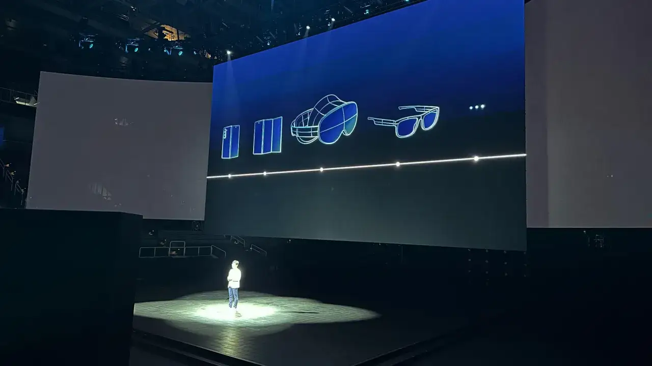 En person st&aring;r p&aring; scenen under en presentation. P&aring; den stora sk&auml;rmen visas en illustration av en vikbar telefon, ett VR-headset och glas&ouml;gon, som en **galaxy unpacked**.