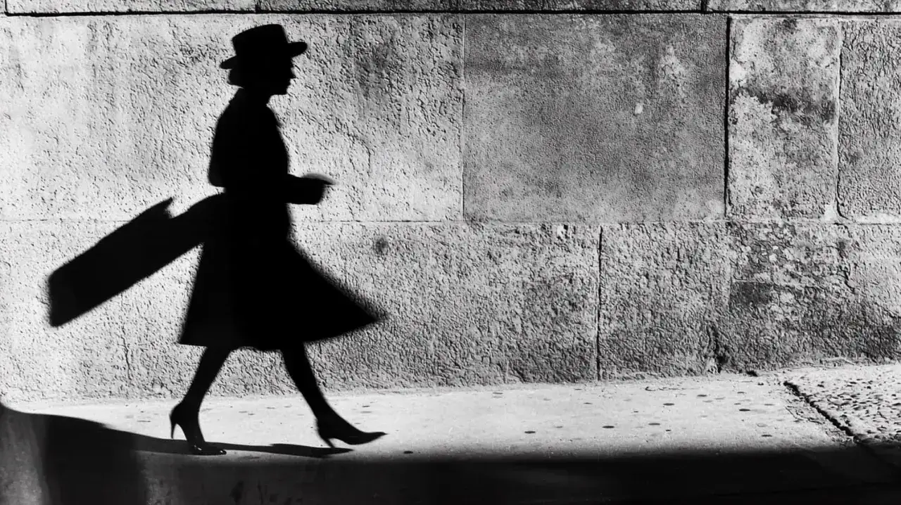 Elegancka kobieta w kapeluszu i płaszczu, uchwycona przez elliott erwitt, przemierza ulicę, rzucając długi, dramatyczny cień.