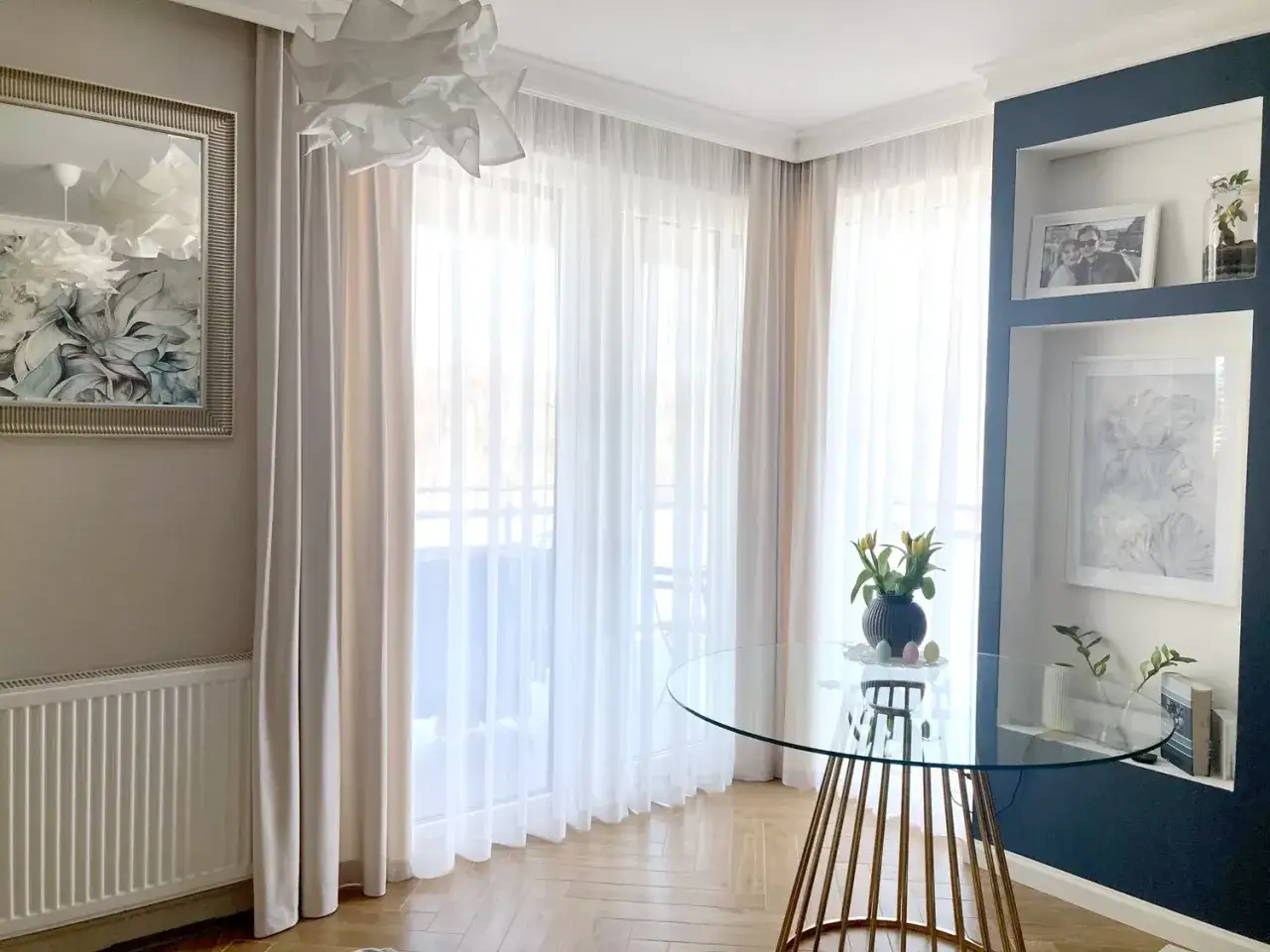 Jasne, świeżo wyprane firanki tworzą przytulną atmosferę w salonie. Na szklanym stole bukiet żółtych tulipanów.