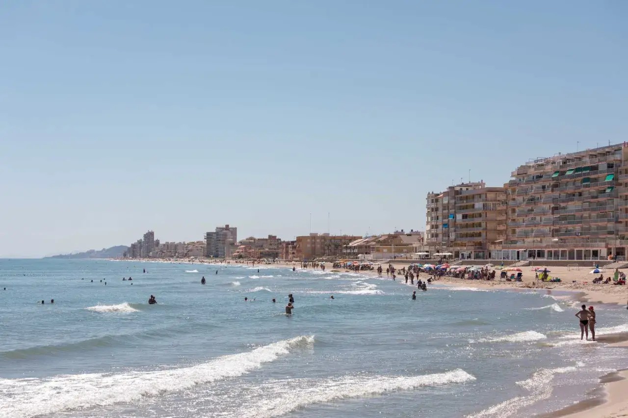 Gente disfrutando del sol y el mar en la playa del Perell&oacute;, con edificios residenciales al fondo.