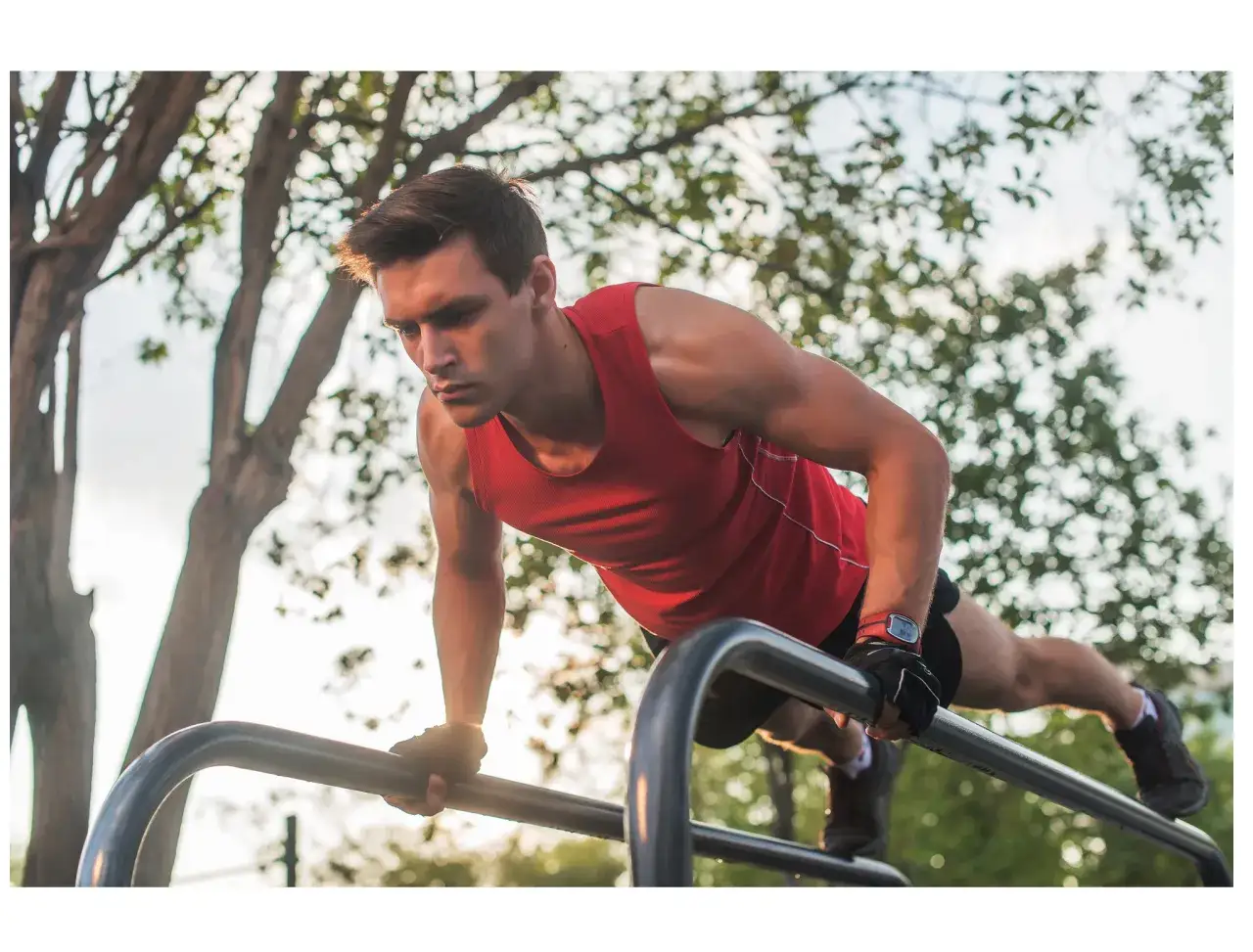 Un homme en d&eacute;bardeur rouge fait des pompes sur des barres de street workout, concentr&eacute; et d&eacute;termin&eacute;.