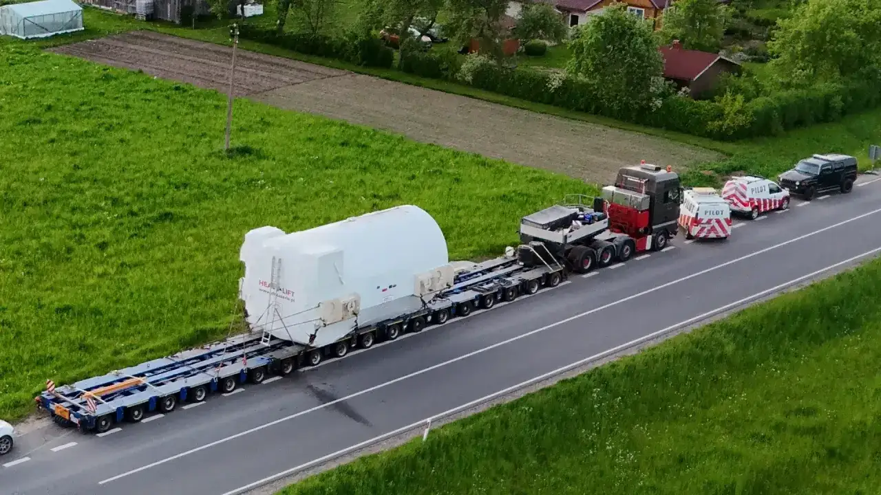 Ogromny ładunek, biały walec, na wieloosiowej platformie. Transport ponadgabarytowy eskortowany przez pojazdy pilotujące.