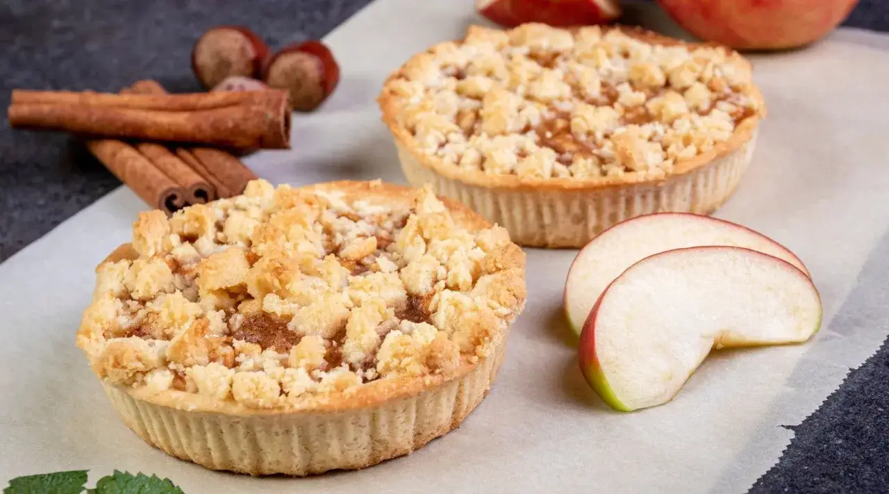 Zwei goldbraune Apfel-Crumble-Törtchen mit Zimtstangen und Äpfeln im Hintergrund.
