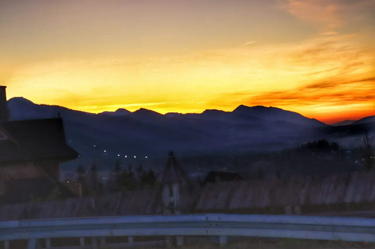 Wschód słońca Zakopane. Góry w odcieniach fioletu i pomarańczy, niebo rozświetlone złotem. Na pierwszym planie drewniany płot i fragment dachu.