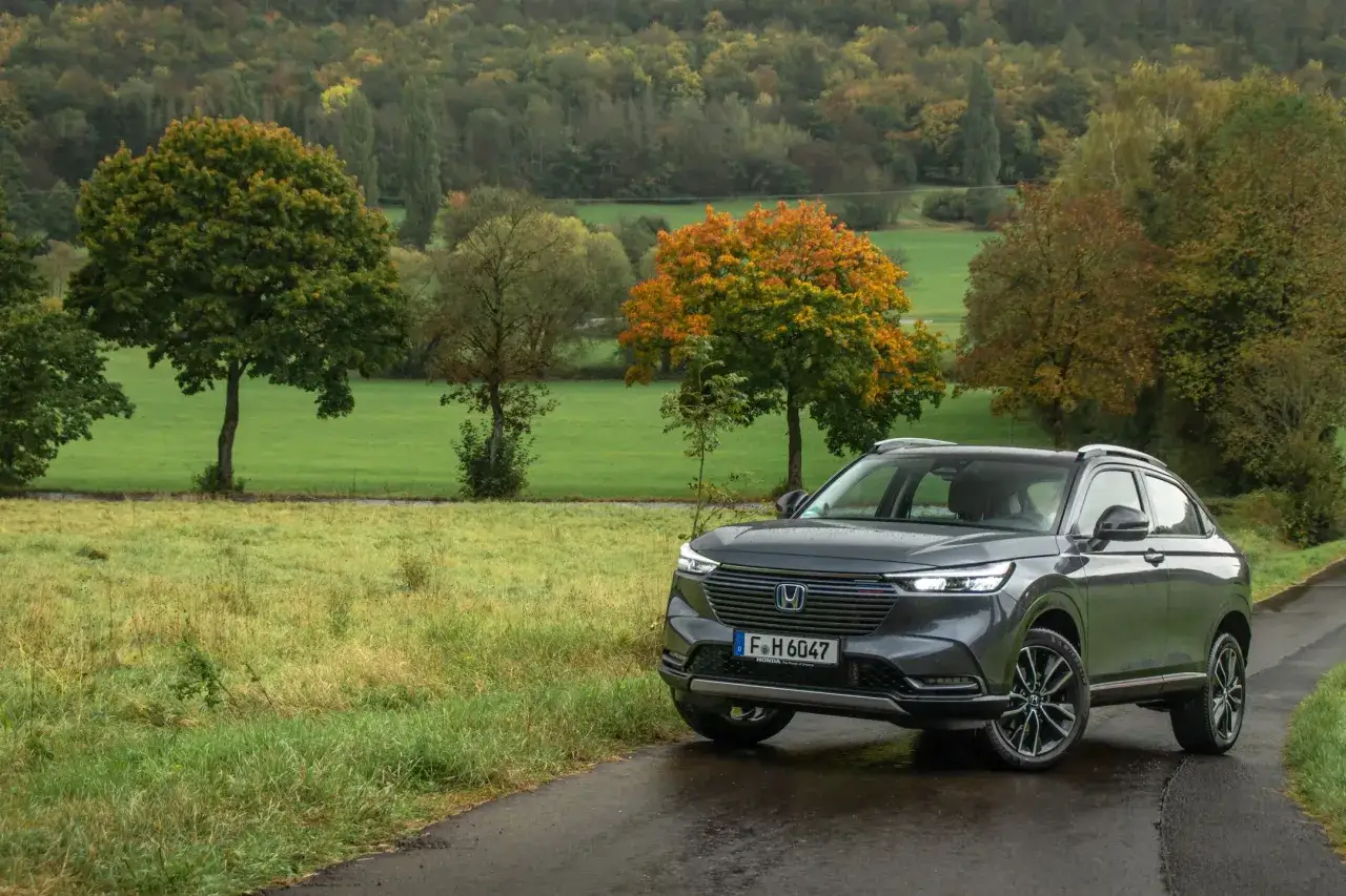 Szary Honda HR-V na mokrej drodze, otoczony jesiennym krajobrazem z zielonymi polami i drzewami w odcieniach żółci i pomarańczy.