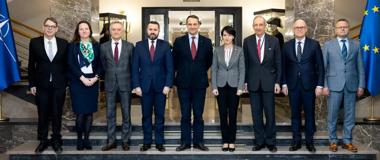 Grupa dyplomatów, w tym minister spraw zagranicznych, pozuje do zdjęcia na tle flag NATO i UE.