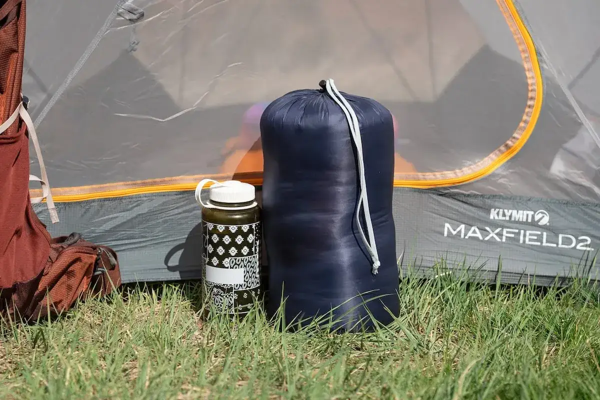 A dark blue sleeping bag, likely a rei magma 15 weight, stands next to a patterned water bottle in front of a Klymit Maxfield2 tent.