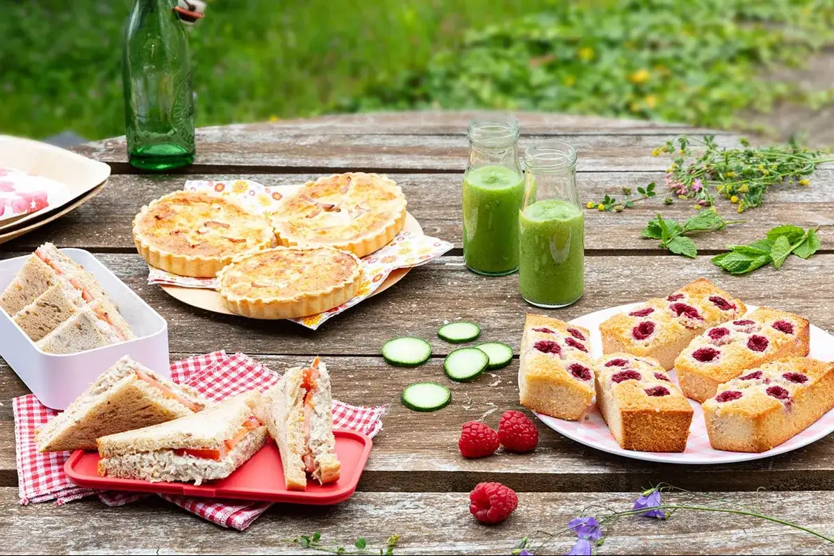 Un délicieux dessert pique-nique : mini-quiches, sandwichs, gâteaux aux framboises et smoothies verts sur une table en bois.