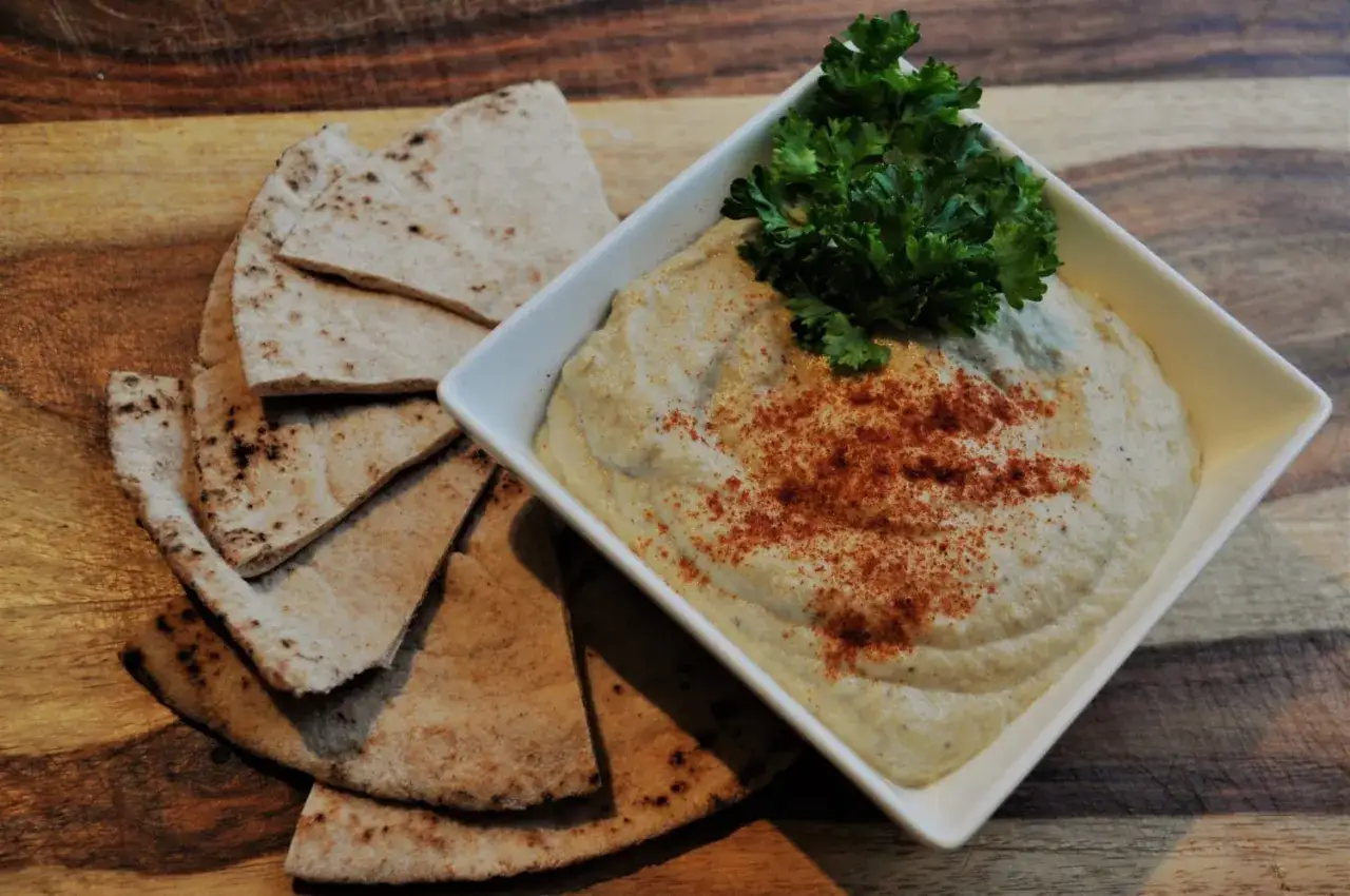 En skål med krämig baba ganoush, toppad med persilja och paprikapulver, bredvid pitabröd på en träskärbräda.