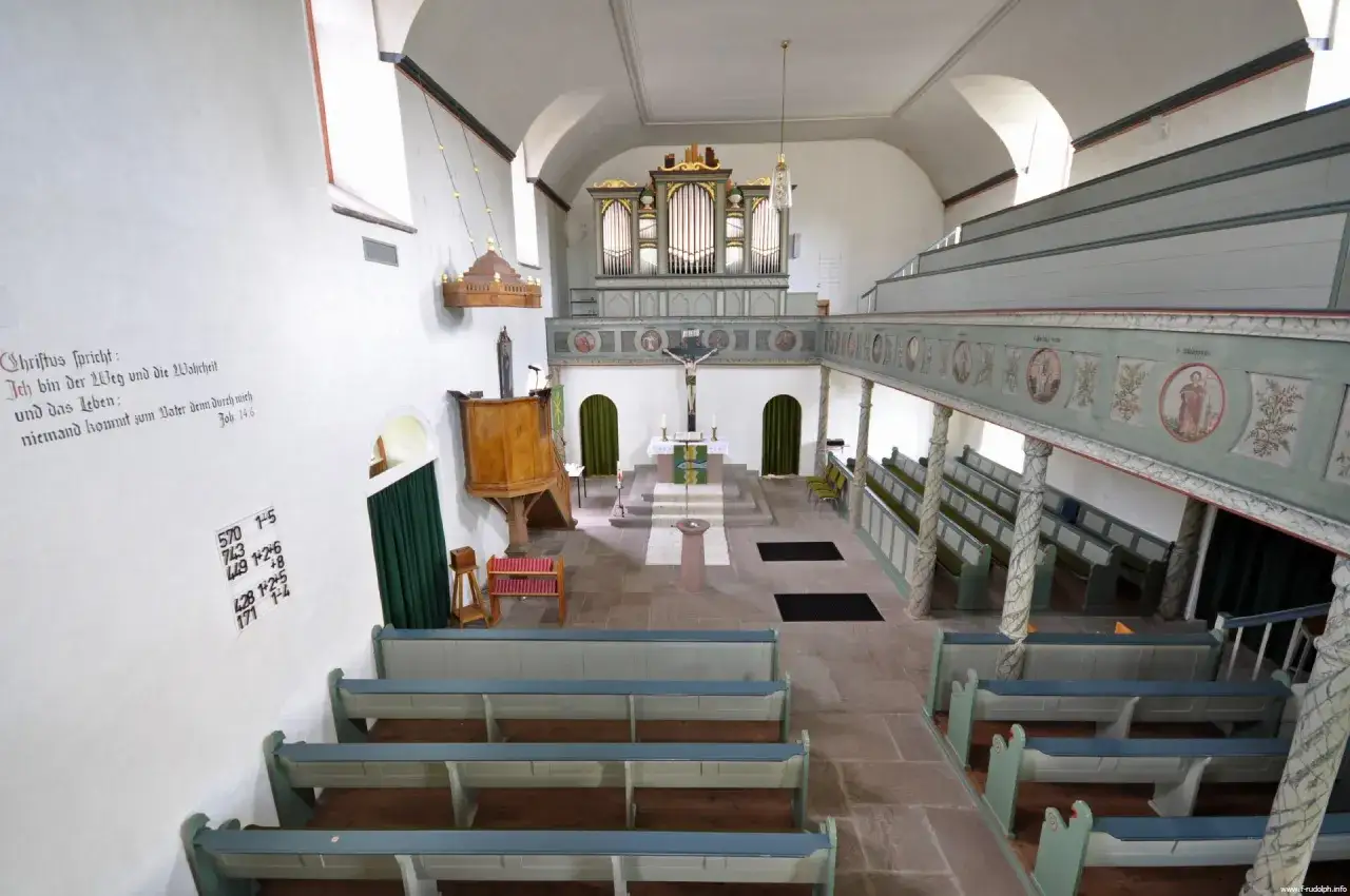 Der Aufbau der evangelischen Kirche zeigt eine Orgel, Kanzel und Emporen mit Bänken. Ein Bibelvers ziert die Wand.