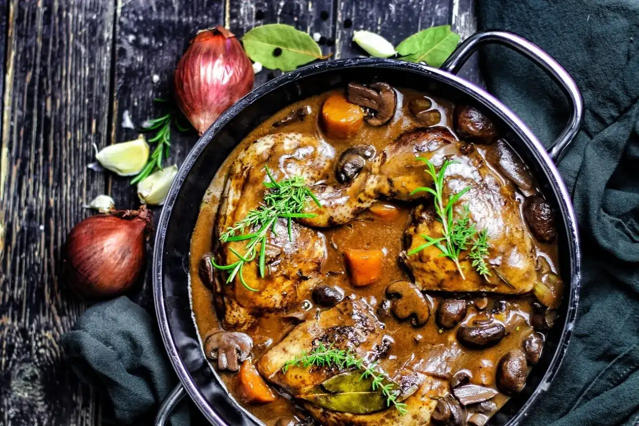 Herzhaftes Coq au Vin in einem Gusseisentopf, mit Pilzen, Karotten und Kräutern garniert. Daneben Zwiebeln und Knoblauch.