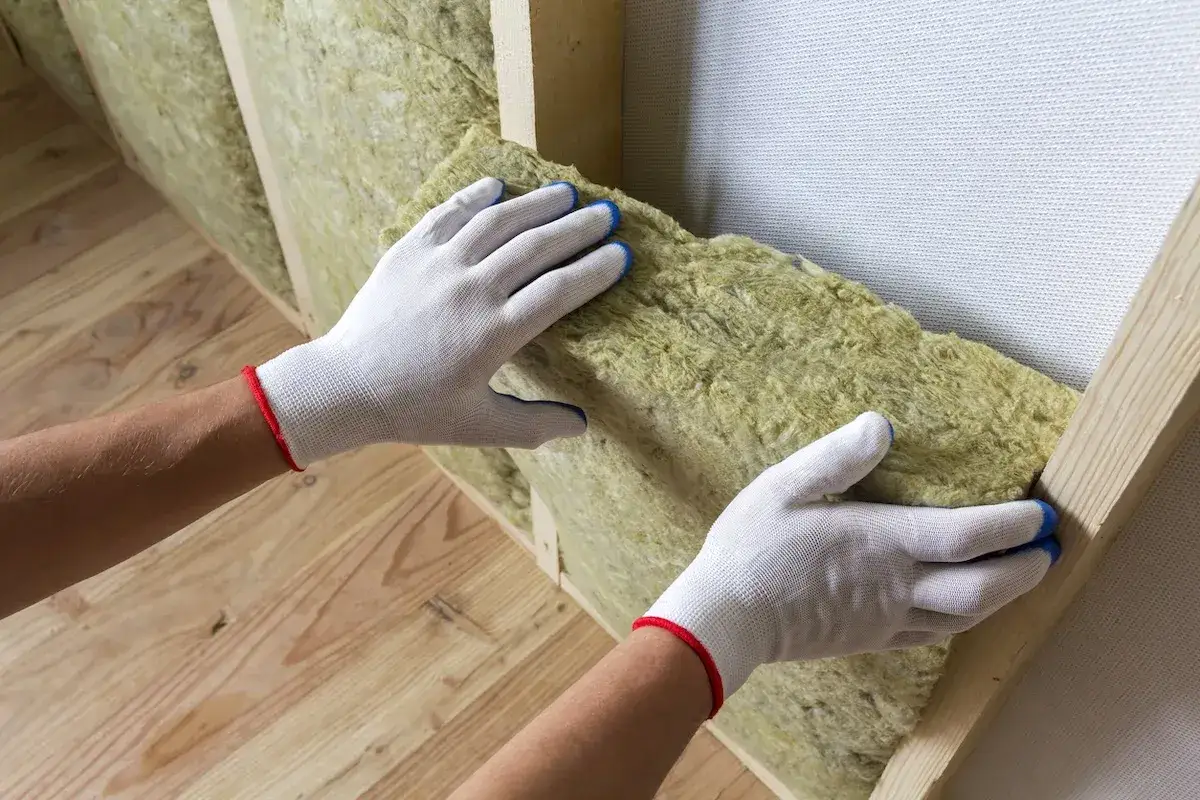 Mani guantate posizionano materiale isolante per un cappotto interno fai da te. Legno e isolamento creano un ambiente caldo.