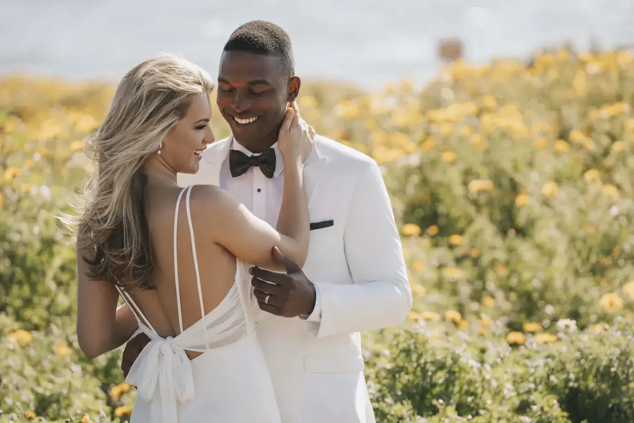 Un couple heureux dans un champ de fleurs jaunes. La mari&eacute;e porte une robe de mariage blanche avec un dos nu. Le mari&eacute; porte un costume blanc.