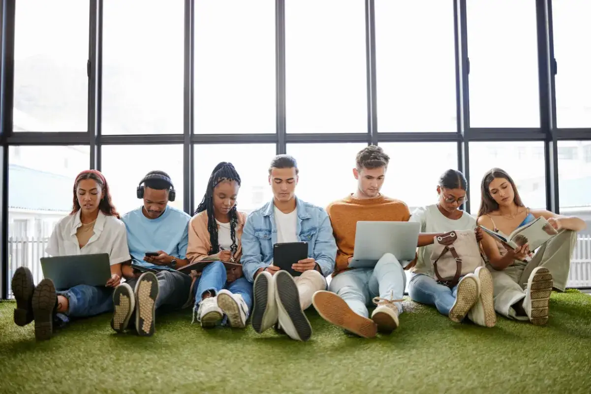 Sechs junge Leute, der **gen z jahrgang**, sitzen auf Kunstrasen vor einem Fenster und arbeiten an Laptops, Tablets oder lesen.