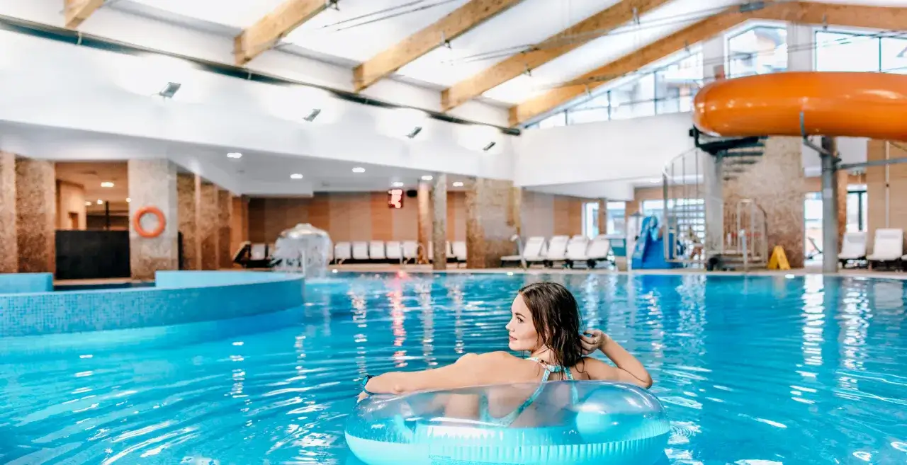 Kobieta relaksuje się w basenie w hotelu z aquaparkiem w górach. Widoczne zjeżdżalnie i leżaki.